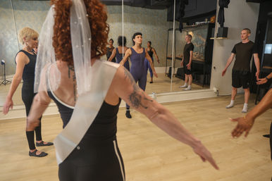 Group dance class in a mirrored studio doing arm warm-ups, led by an instructor in a purple leotard, with a foreground participant wearing a veil and sash