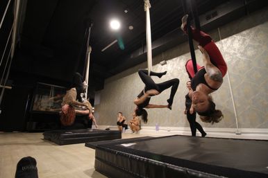 Aerial silks class in a studio: students suspended upside-down from hanging white silks over padded crash mats, practicing inverted poses against patterned wallpaper and exposed black ceiling.