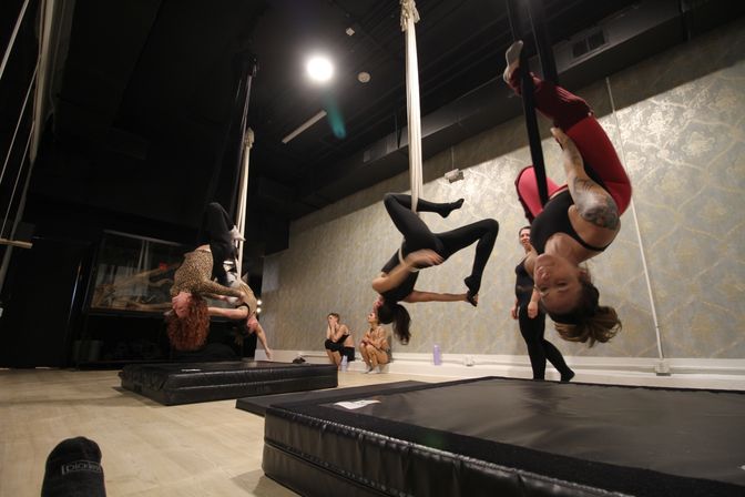 Aerial silks class in a studio: students suspended upside-down from hanging white silks over padded crash mats, practicing inverted poses against patterned wallpaper and exposed black ceiling.