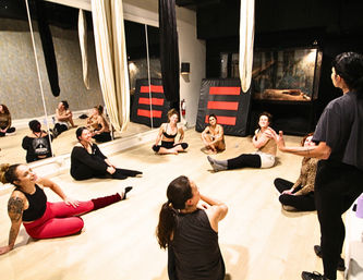 Group aerial-silks class in an indoor fitness studio: participants sit in a circle on a light wood floor, laughing and stretching beneath hanging fabric silks with mirrored walls in the background.