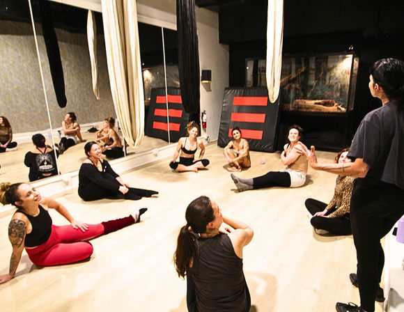 Group aerial-silks class in an indoor fitness studio: participants sit in a circle on a light wood floor, laughing and stretching beneath hanging fabric silks with mirrored walls in the background.