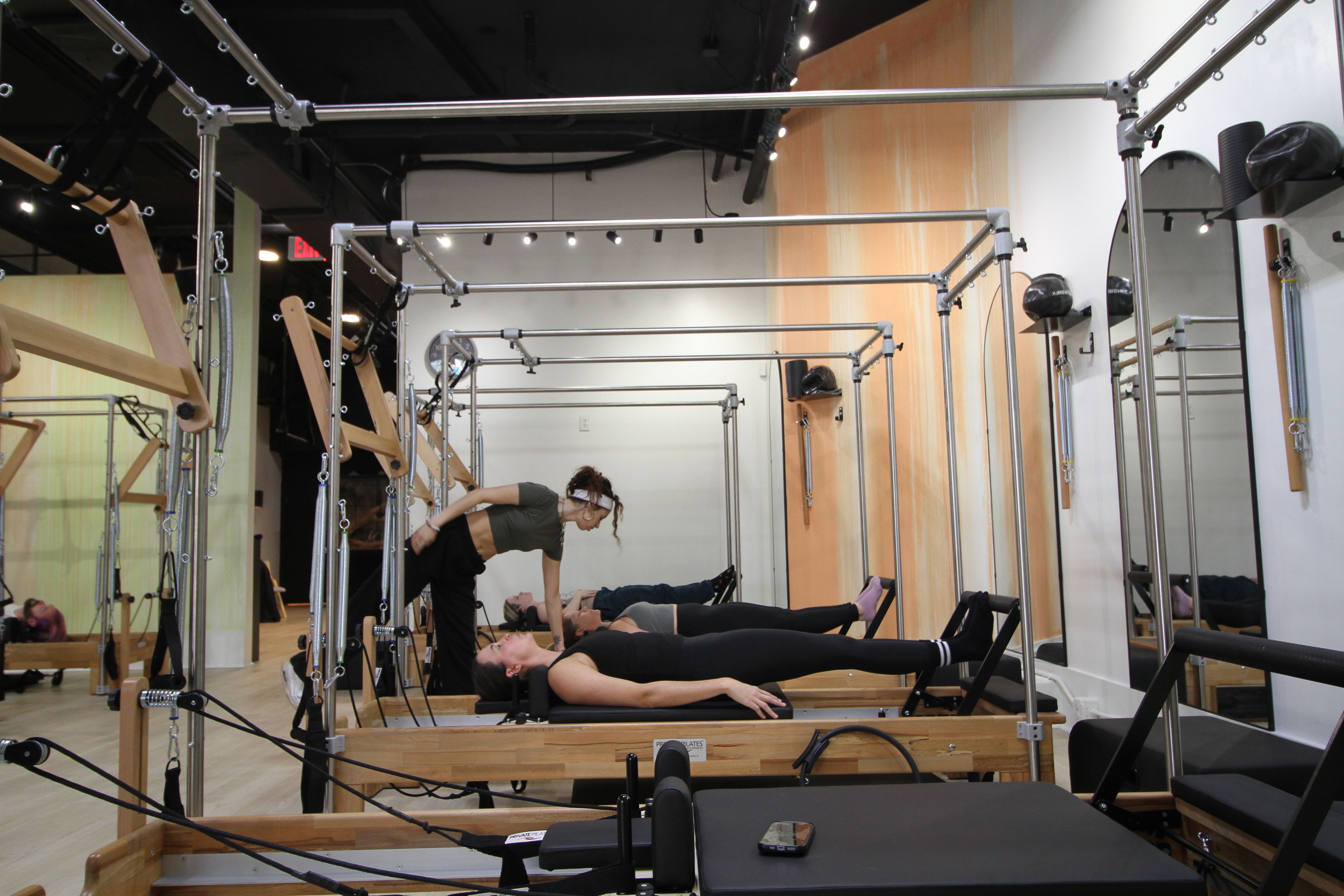 Instructor adjusting a student on a wooden Pilates reformer during a workout in a bright mirrored fitness studio.