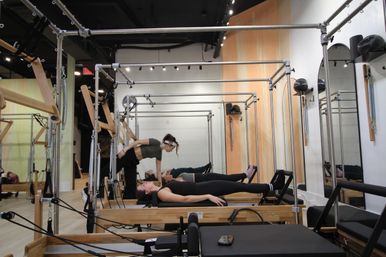 Instructor adjusting a student on a wooden Pilates reformer during a workout in a bright mirrored fitness studio.