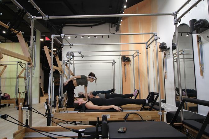 Instructor adjusting a student on a wooden Pilates reformer during a workout in a bright mirrored fitness studio.