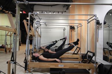Boutique Pilates studio interior with wooden reformers in a row; two participants lie supine on carriages using straps to lift their legs, mirrored wall and light wood floors.