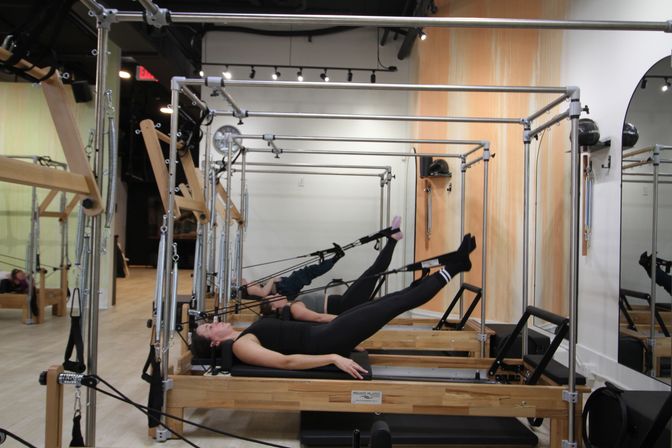 Boutique Pilates studio interior with wooden reformers in a row; two participants lie supine on carriages using straps to lift their legs, mirrored wall and light wood floors.