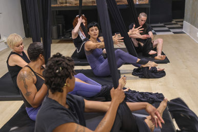Group aerial yoga class in a fitness studio — instructor demonstrating a seated hammock pose while diverse students sit on mats holding black aerial silks