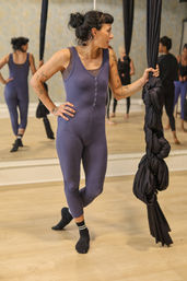 Tattooed woman in a purple jumpsuit grips knotted black aerial silks in a mirrored fitness studio during an aerial silks class.