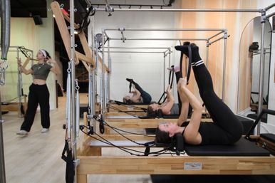 Pilates reformer class in a bright fitness studio with participants lying on reformers doing leg stretches while an instructor moves in the background