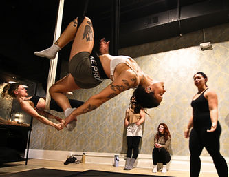 Dynamic indoor fitness studio scene: women practicing pole and aerial silks, two participants suspended upside down in a synchronized inverted pole move while classmates watch on mats.