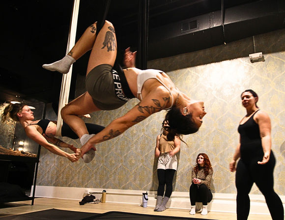 Dynamic indoor fitness studio scene: women practicing pole and aerial silks, two participants suspended upside down in a synchronized inverted pole move while classmates watch on mats.