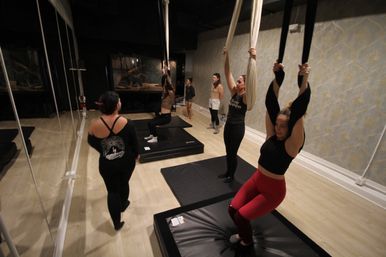 Aerial yoga class in an indoor studio with women practicing on black and white silk hammocks above padded mats, mirrored wall, patterned wallpaper and light wood floor.