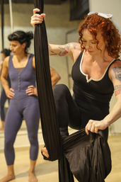 Aerial yoga class in a fitness studio — a red‑haired woman with tattoos adjusting a black aerial silk hammock while another participant watches.