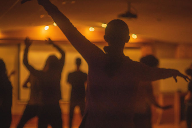 Silhouetted dancers in a warm-lit indoor studio during a lively group dance class.