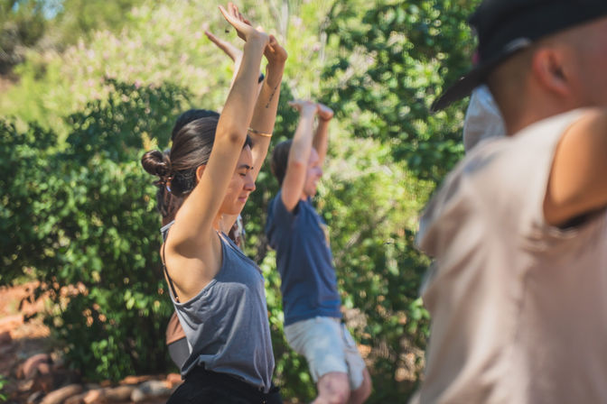 Group practicing yoga outdoors in a sunlit park, standing with arms raised amid lush green trees and shrubs.