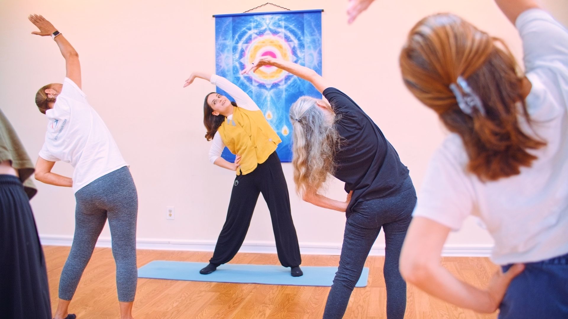 Adults in a bright yoga studio doing synchronized side-bend stretches on mats beneath a colorful mandala tapestry.