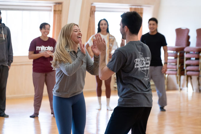 Smiling young adults in a bright community dance studio doing a partner exercise — a woman and man playfully high-five while others watch on a polished wooden floor during a group dance workshop.