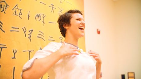 Joyful person with short hair speaking or singing animatedly in a bright art gallery, standing before a large yellow wall covered in black ancient-style symbols.
