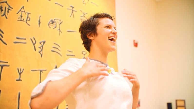 Joyful person with short hair speaking or singing animatedly in a bright art gallery, standing before a large yellow wall covered in black ancient-style symbols.