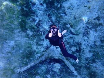 Underwater snorkeler in a black wetsuit and mask giving double thumbs-up while floating above a rocky, sandy seafloor in clear blue water.