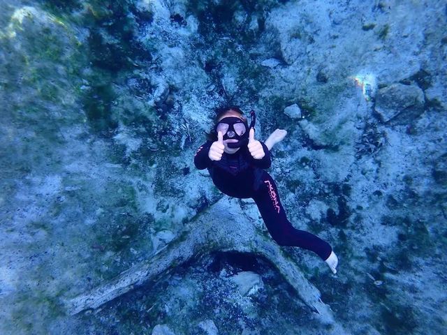 Underwater snorkeler in a black wetsuit and mask giving double thumbs-up while floating above a rocky, sandy seafloor in clear blue water.