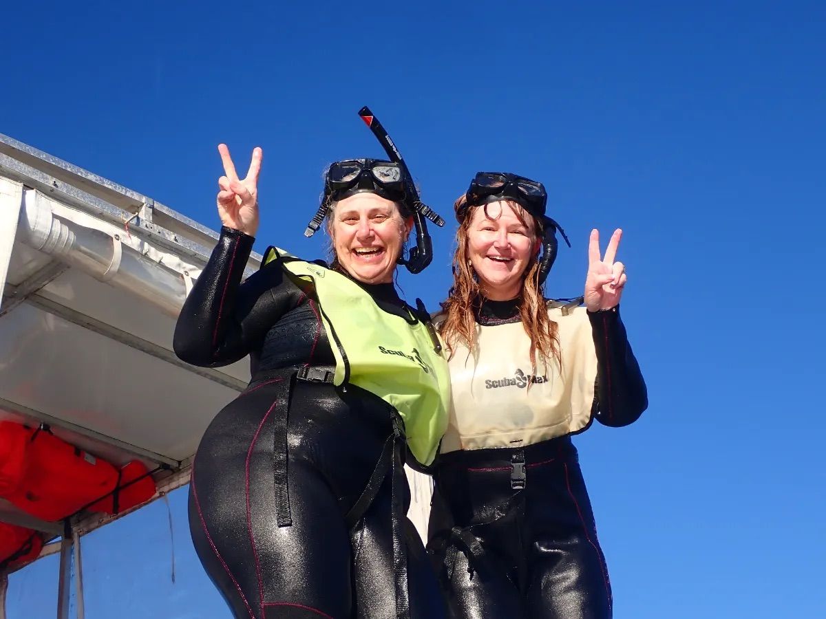 Two cheerful snorkelers in black wetsuits and snorkel masks flashing peace signs on a boat deck against a clear blue sky