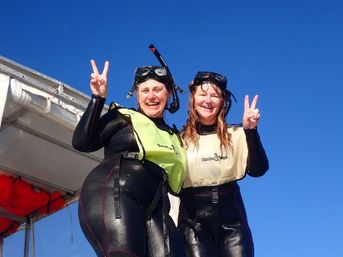 Two cheerful snorkelers in black wetsuits and snorkel masks flashing peace signs on a boat deck against a clear blue sky