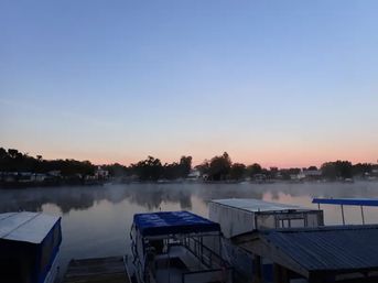Sunrise over a calm lake with pastel sky, thin morning mist on the water, tree-lined shoreline and covered boats docked at a small marina