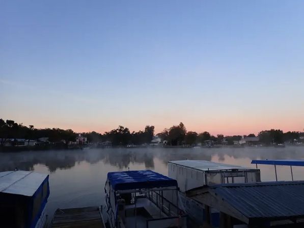 Sunrise over a calm lake with pastel sky, thin morning mist on the water, tree-lined shoreline and covered boats docked at a small marina