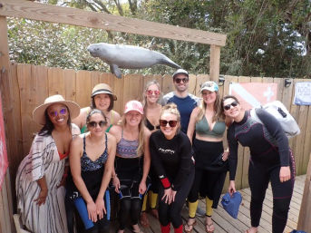 Smiling group in wetsuits and beachwear posing on a wooden dock beneath a hanging manatee sculpture, ready for a coastal wildlife tour.