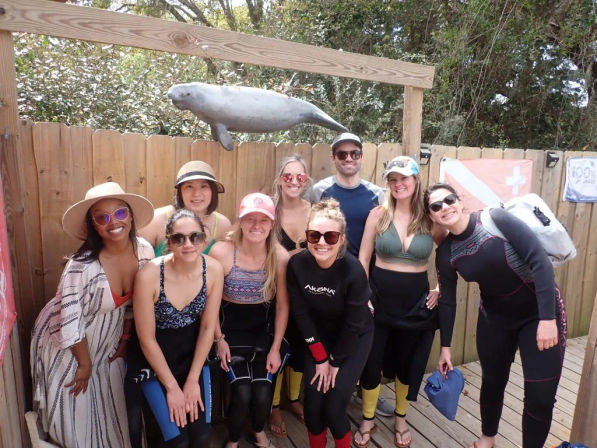 Smiling group in wetsuits and beachwear posing on a wooden dock beneath a hanging manatee sculpture, ready for a coastal wildlife tour.