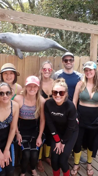 Smiling group in wetsuits and beachwear posing on a wooden dock beneath a hanging manatee sculpture, ready for a coastal wildlife tour.
