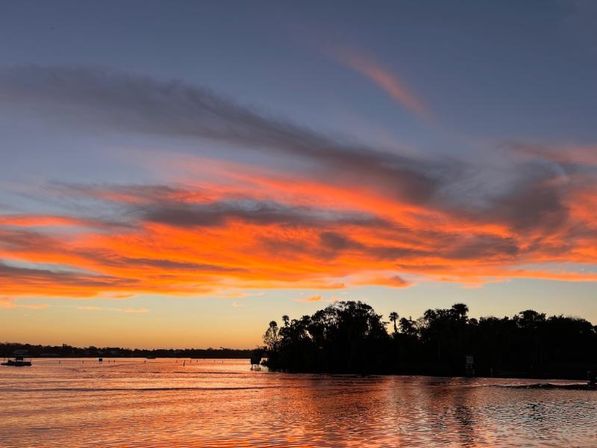 Vibrant orange and pink sunset with dramatic cloud streaks reflecting on calm bay waters and a silhouetted tree-covered island on the horizon