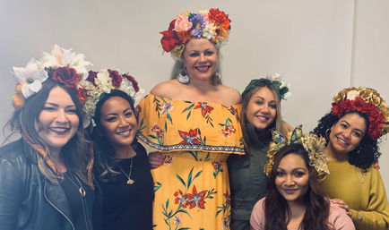 Six smiling women in an indoor group portrait wearing colorful floral crowns, center woman in a yellow off-shoulder floral dress, festive celebration vibe.