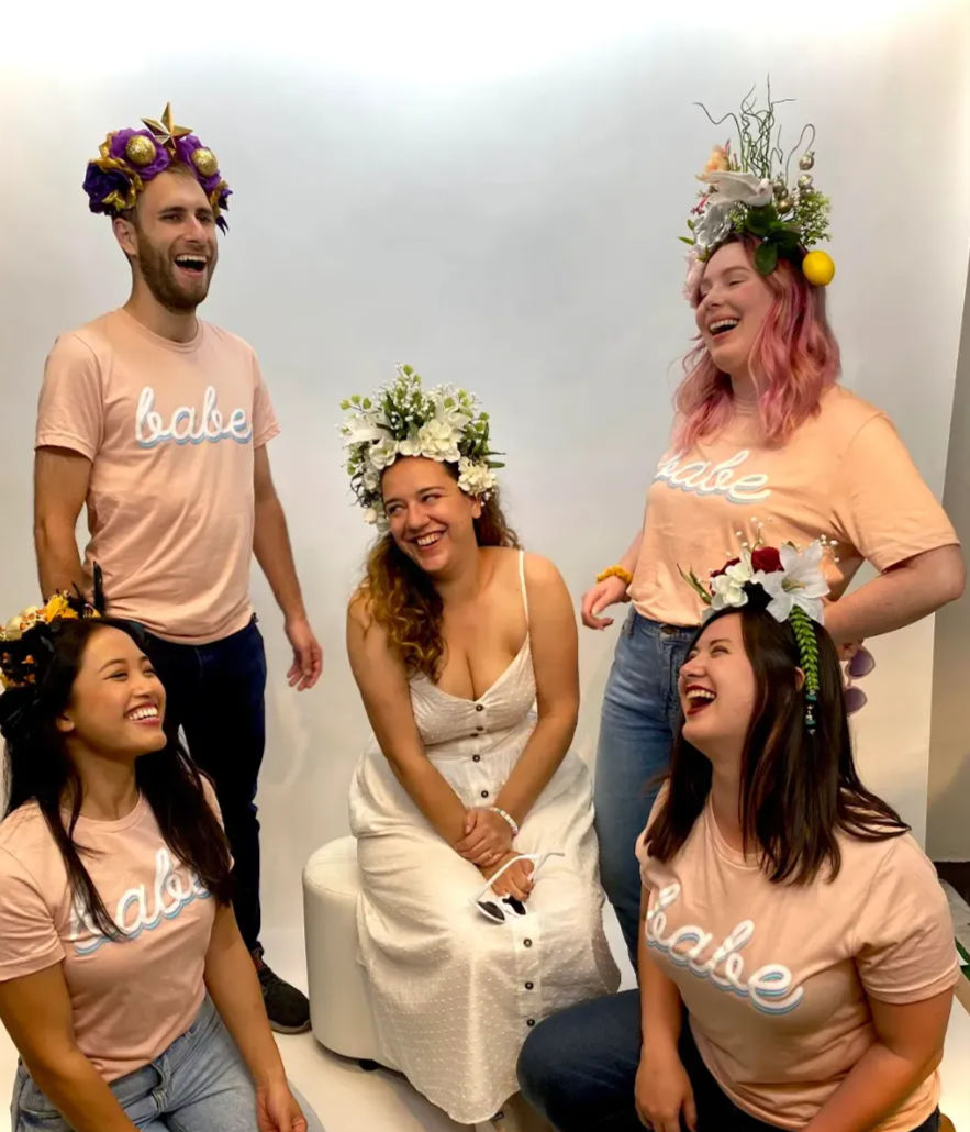 Smiling group portrait of five friends wearing colorful floral crowns; four in matching peach 'babe' tees and one seated in a white sundress, laughing against a plain studio backdrop.