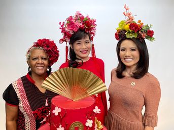 Three smiling women wearing colorful floral headpieces pose behind a tall red cake adorned with cherry blossoms and a large gold fan topper, festive Lunar New Year–inspired scene.