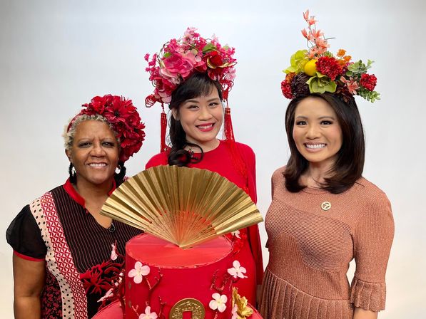Three smiling women wearing colorful floral headpieces pose behind a tall red cake adorned with cherry blossoms and a large gold fan topper, festive Lunar New Year–inspired scene.