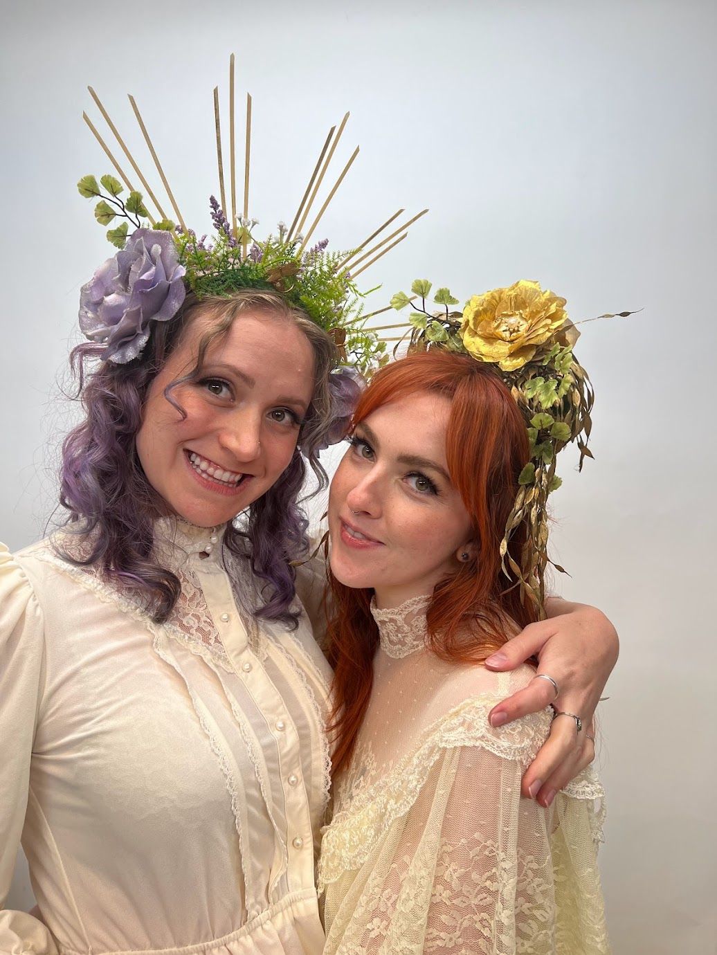 Two women in cream lace dresses pose in a studio, smiling while wearing floral halo headpieces—one with lavender hair and a purple flower, the other with red hair and a golden rose—whimsical vintage-inspired portrait