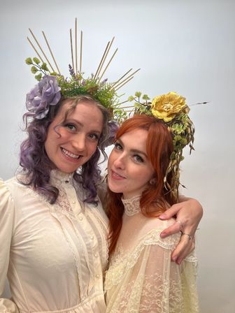 Two women in cream lace dresses pose in a studio, smiling while wearing floral halo headpieces—one with lavender hair and a purple flower, the other with red hair and a golden rose—whimsical vintage-inspired portrait