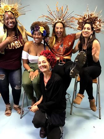 Five women wearing ornate floral and sunburst headpieces smiling and posing in a light-blue photo studio, seated on stools and kneeling for a playful group portrait.