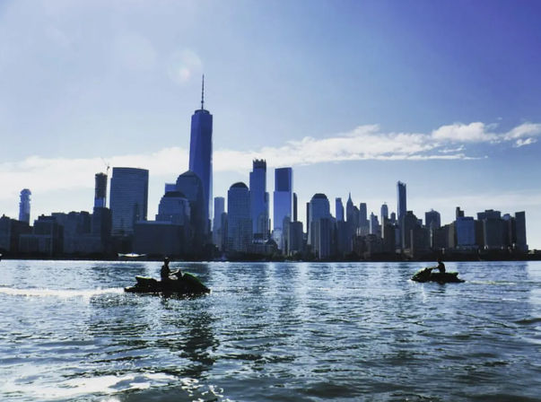 Two jet-ski riders zip across the Hudson River with New York City skyline, including One World Trade Center, shimmering under a bright blue sky