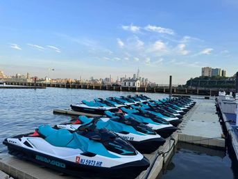 Fleet of teal-and-white personal watercraft lined up on a floating dock along the Hudson River with the Lower Manhattan skyline and One World Trade Center in the distance under a clear blue sky.