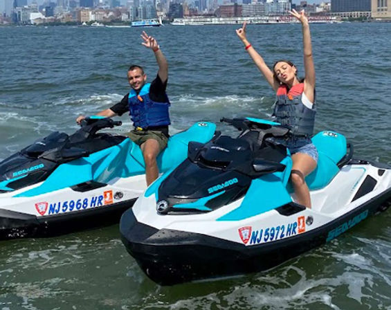 Cheerful pair in life vests on matching turquoise-and-white jet skis, waving as they ride through a city harbor with the skyline in the background.