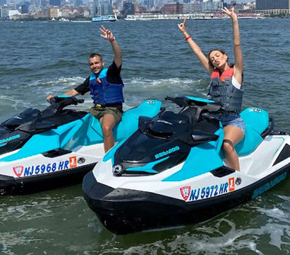 Cheerful pair in life vests on matching turquoise-and-white jet skis, waving as they ride through a city harbor with the skyline in the background.
