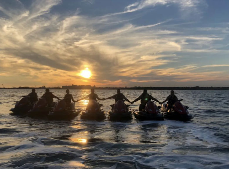 Seven riders on jet skis holding hands in silhouette at sunset over open water, dramatic cloud-streaked sky and golden reflections on rippled waves.