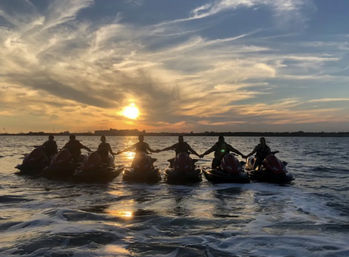 Seven riders on jet skis holding hands in silhouette at sunset over open water, dramatic cloud-streaked sky and golden reflections on rippled waves.
