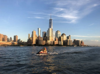 Two riders on a jet ski crossing the Hudson River with Lower Manhattan skyline and One World Trade Center glowing at golden hour