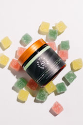 Flat-lay product photo: amber-lidded jar of gummy supplements surrounded by green, yellow, and pink sugar-coated square gummies on a white background.