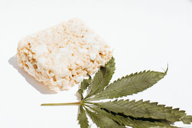 Rice cereal marshmallow square beside a cannabis leaf on a bright white background — playful edible/hemp concept.