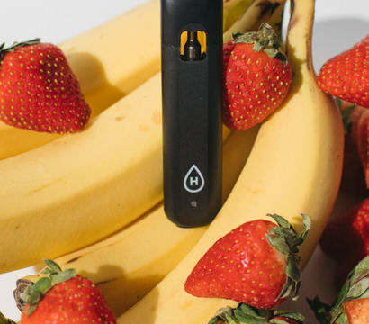 Playful close-up of a black vape pod device nestled among ripe yellow bananas and red strawberries on a bright white background — a summer fruit still-life composition.